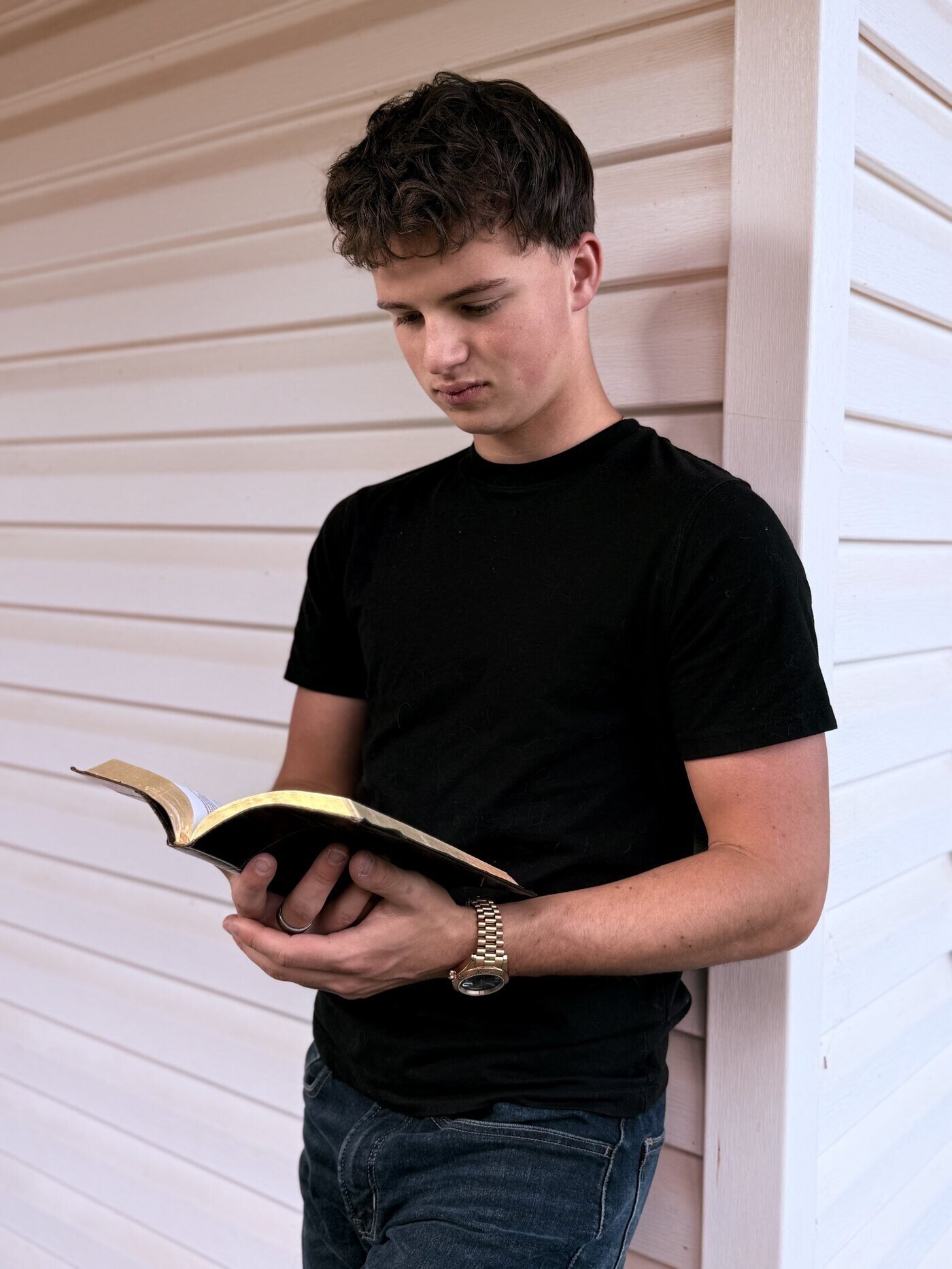 Jake leaning against a white clapboard wall, reading an open Bible.